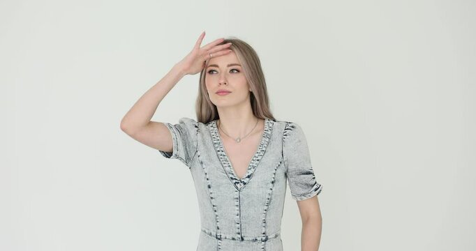 Blonde woman raises hand to forehead on gray background. Lady shields from sun squinting in distance searching intently for someone in bright light