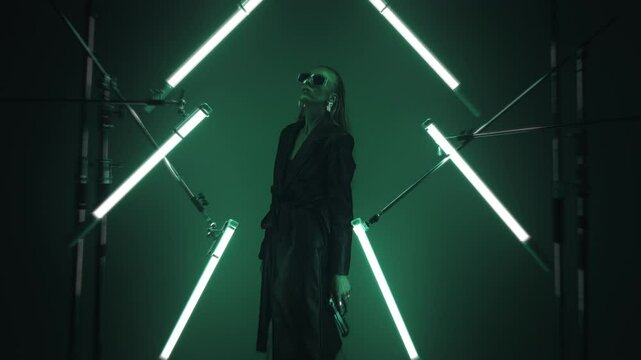 Beautiful futuristic lady in leather coat and sunglasses holding a gun and posing in a dark room lit by RGB tube lights