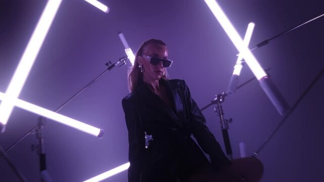 Gorgeous elegant female secret agent in high heels and leather coat holding a gun while sitting in a dark studio room surrounded by rgb tube lights