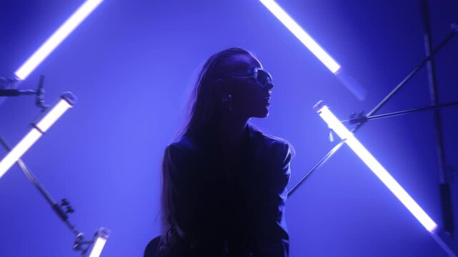 Beautiful futuristic lady in leather coat and sunglasses holding a gun and posing in a dark room lit by RGB tube lights