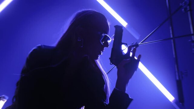 Beautiful futuristic lady in leather coat and sunglasses holding a gun and posing in a dark room lit by RGB tube lights