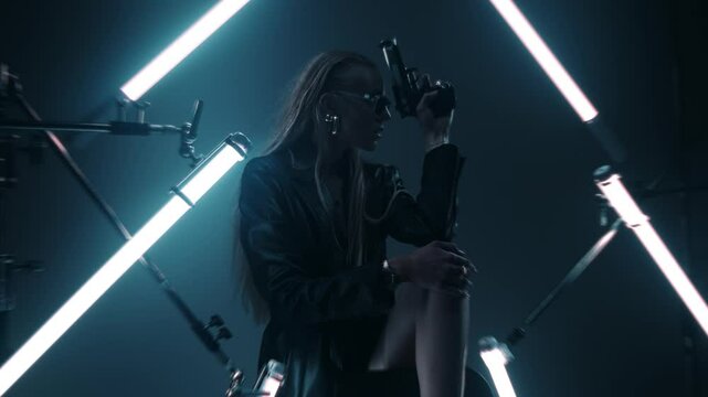 Beautiful futuristic lady in leather coat and sunglasses holding a gun and posing in a dark room lit by RGB tube lights