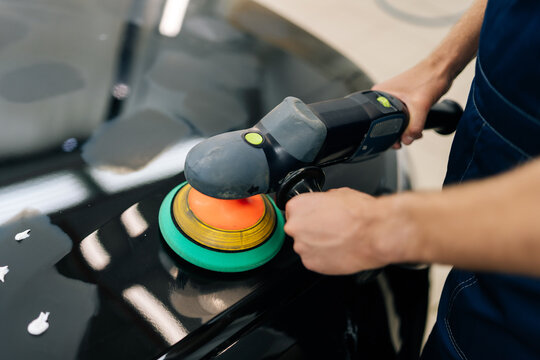 Closeup top view of unrecognizable mechanic male in protective gloves polishing surface of hood of black car using orbital polisher in repair shop. Concept of professional car repair, maintenance work - Powered by Adobe