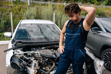 Confused mechanic scratching head in frustration while assessing damage on car after an accident. Vehicle service repairman looking car front end completely destroyed in garage car service.