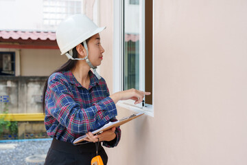 Asian female construction inspector in white hard hat pointing at building window during an outdoor inspection. Wearing a plaid shirt and holding a clipboard, observing details on the window.