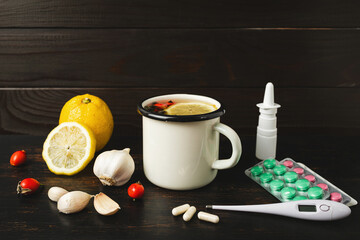 Pharmacy medicine and natural remedies, thermometer, white capsules, pills in blister pack, nasal spray and cup of herbal tea, lemon, garlic, ginger and dog rose fruit on a dark wooden background