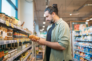 A young man is shopping at the supermarket. The guy picks canned food from the counter in the store