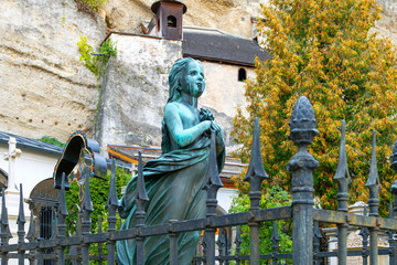 Naklejka premium Bronze statue of a young girl as a memorial headstone at a grave in the St Peter's Cemetery, with the ancient abbey Catacombs behind, in Salzburg, Austria.
