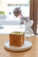Close-up side view of freshly baked brown cube croissant with green pistachio cream on wooden table by flower in vase in restaurant or cafe. Soft focus. Copy space. Puff pastry theme.