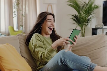 Obraz premium Joyful young woman enjoying online shopping on her smartphone while relaxing on a cozy couch in her home during a sunny afternoon