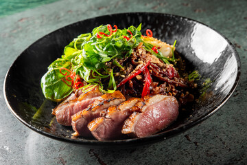 gourmet dish featuring sliced duck breast, fresh greens, and chili garnish, elegantly presented in a black bowl on a textured surface. The vibrant colors and textures highlight the culinary artistry.