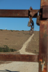 A sunny day with clear skies, there is a rusty metal gate on a field with a rusty lock on it. Being the gate one sees a gravel road going up the hill. 
