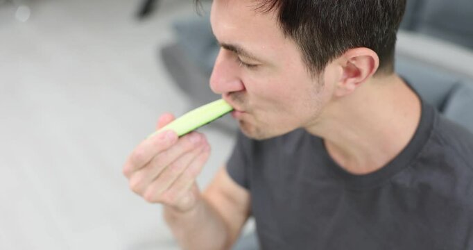 Hungry man bites slice of fresh cucumber sitting on sofa in living room. Guy enjoys ripe vegetable with juicy texture appreciating taste
