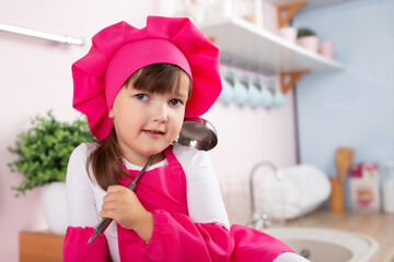 Little beautiful girl in a chef costume prepares food while sitting near the cooker in the kitchen.