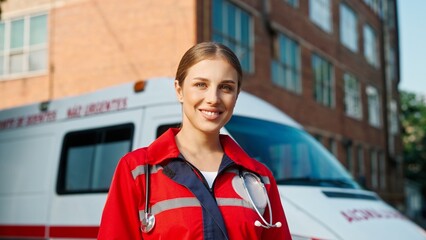 Close up portrait of attractive young female medical worker in red uniform standing on street in front of ambulance car and looking at camera. Positive happy pretty woman outdoors. Healthcare concept © VAKSMANV