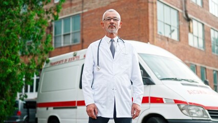 One handsome Caucasian senior experienced professional man doctor in white robe with stethoscope standing outdoors in city looking at camera near ambulance vehicle. Emergency, medical concept © VAKSMANV