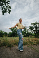 Fototapeta premium Young woman posing confidently outdoors in a casual outfit with ripped jeans and a yellow top, set against a cloudy sky.