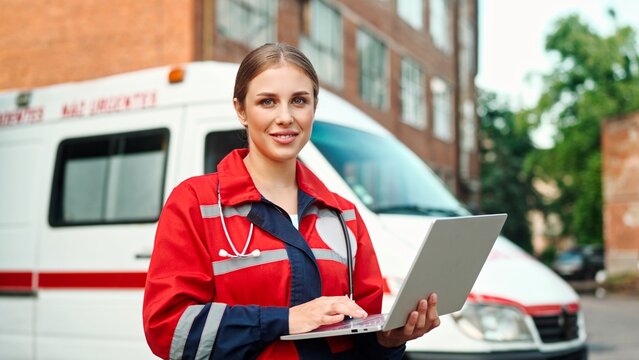Camera moving back from beautiful Caucasian young female medical worker in red uniform typing on laptop standing on street near hospital. Woman paramedic using computer outdoor near ambulance