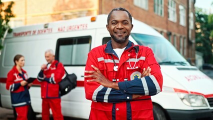 Joyful smiling African American male doctor in red medical uniform looking at camera standing outdoor near ambulance vehicle. Paramedics with tablet device on background, first aid, healthcare concept