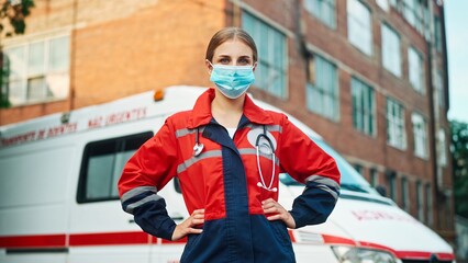 Obraz premium One young beautiful woman paramedic standing outdoor wearing medical mask and looking at camera near ambulance vehicle. Emergency worker. Professional doctor or nurse at work. First aid assistance