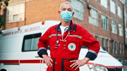 One Caucasian senior experienced man paramedic standing outdoor wearing medical mask on face and looking at camera near ambulance vehicle. Emergency worker. Professional doctor. First aid assistance © VAKSMANV