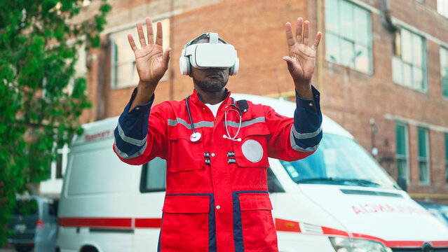 Portrait of African American male medical worker in VR headset making gestures with hands in air standing outdoors near ambulance. Futuristic. Professional paramedic. Virtual reality, metaverse - Powered by Adobe