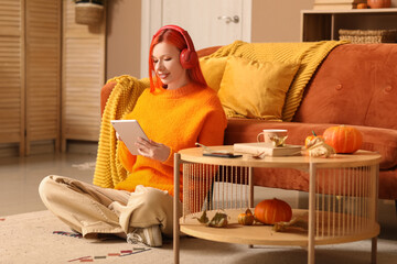 Beautiful young happy woman in headphones with autumn decor and tablet sitting on floor at home