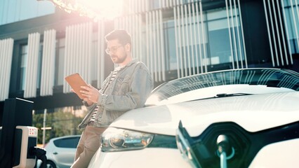 Young joyful handsome man typing on tablet device while waiting till his electric car charge. Male...