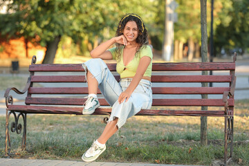 Happy young African-American woman in headphones sitting on bench in park