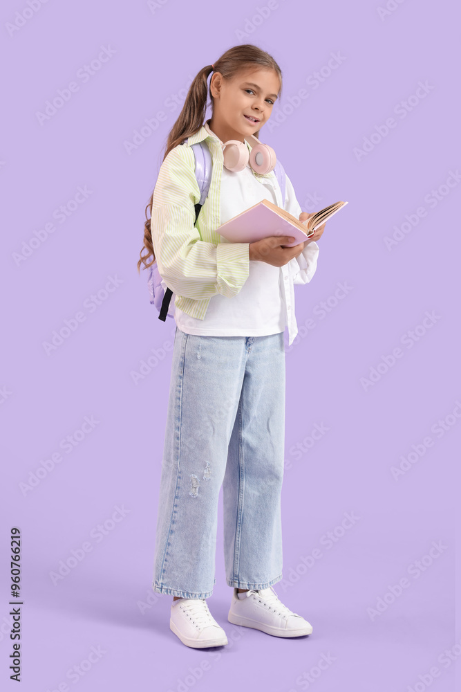 Wall mural Cute schoolgirl reading book with headphones and backpack on lilac background