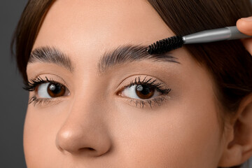 Young woman correcting shape of her eyebrows on grey background, closeup