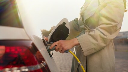 Happy young woman using city charger station to charge her auto on street. Female charging EV. Electric vehicle. Caucasian pretty woman charges electric car at charger point. Technology, close up