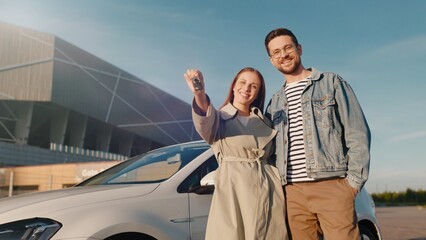 Happy cute Caucasian couple hugging and smiling outdoor near new electric car. Cheerful man and...