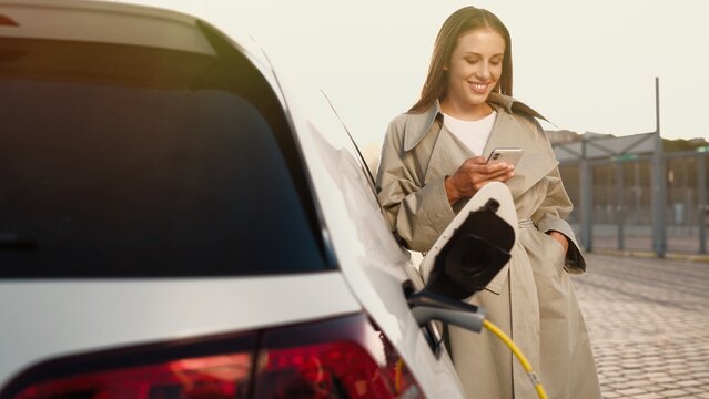 Cheerful attractive stylish woman smiling while tapping on cell phone standing on street waiting for her electric car to charge. Happy female texting on smartphone charging EV, electrocar concept
