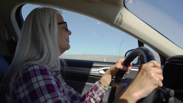Old woman drive a car in hot and sunny day on highway. A woman with glasses is driving a car at deserts area during sunshine. Uber driver driving in beautiful place