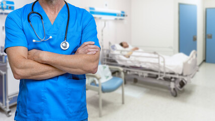 Nurse in blue scrubs with arms crossed in a hospital room with an ill patient in bed.