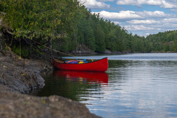 Bright red canoe loaded with camping gear tied to a tree by rugged rocky shore at French River...