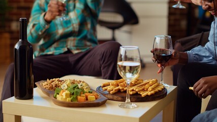 Close up shot of charcuterie board on stylish living room table next to group of friends talking. Tasty gourmet cheese platter ready to be enjoyed by apartment party guests