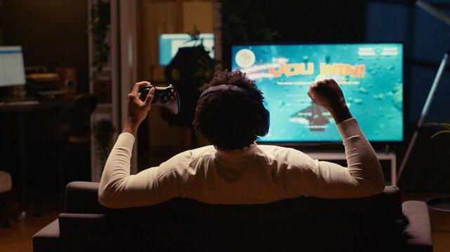 BIPOC man happy after winning SF game, destroying all enemy spacecrafts and finishing level. Player feeling excited after victory in videogame displayed on widescreen TV, defeating boss to save galaxy