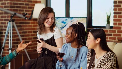 Obraz premium Best friends laughing together, enjoying alcohol beverages and gourmet cheese platter in cozy apartment living room. BIPOC group of people having lively conversation at home, entertaining each other