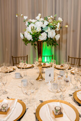 Round table in a restaurant decorated with white roses and empty golden plates waiting for guests. Cutlery and golden plates on a table in a restaurant. Vertical photo of the table