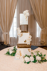 Gorgeous tiered wedding cake decorated with edible gold and white roses. White luxury wedding cake stands on a table in a restaurant. Beautifully decorated cake for the bride and groom