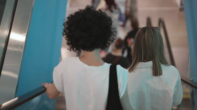 Two women with curly hair standing on moving escalator in airport, train station, shopping mall, cinematic isolated shot