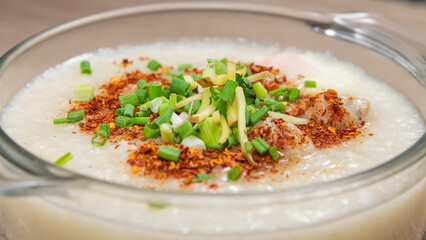 Pork congee with boiled eggs in a glass bowl, topped with sliced ginger, sliced green onions and red chili powder