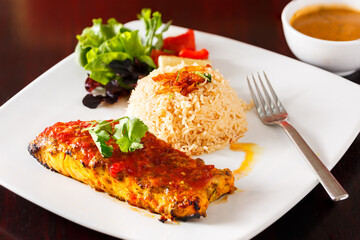 Indian Tandoori Salmon with a mound of rice and salad greens in a white plate on a dark table with condiment on the side; copy space