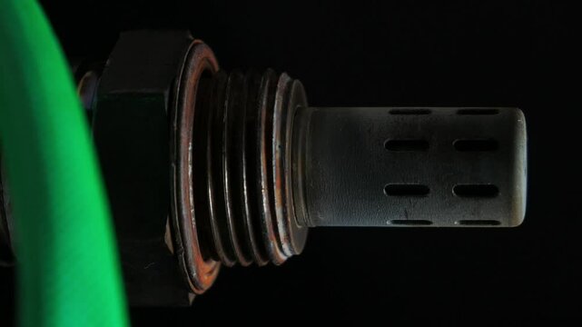 Rusty lambda sensor rotates slowly against a black backdrop. Covered in dirt and grime, the metal shows signs of corrosion and decay, highlighting its weathered condition
