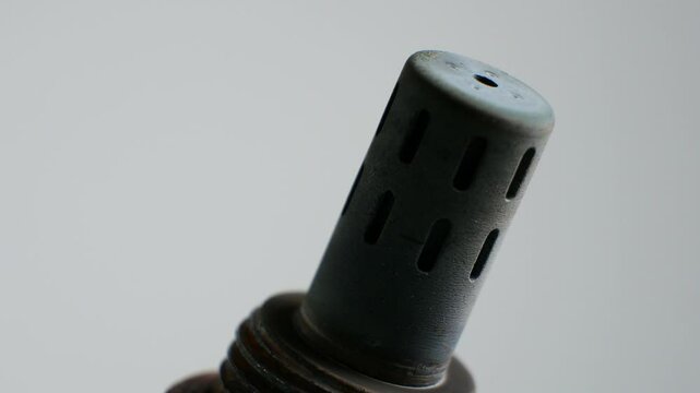 Mechanic rotates a dirty oxygen sensor against a white background. The sensor shows signs of grime and wear, highlighting the importance of regular vehicle maintenance and inspection