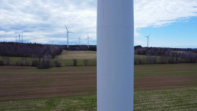 Un drone remonte lentement un mat d'&eacute;olienne jusqu'a son rotor situ&eacute; au sommet avec en arri&egrave;re-plan des champs cultiv&eacute;s, de la for&ecirc;t et d'autres &eacute;oliennes en rotation sous un ciel bleu nuageux.