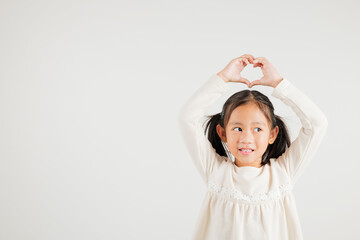 Fototapeta premium A smiling Asian young kid girl shows confidence, creating a heart shape with fingers and hands on white background. Portrait of kindergarten child sending love for Valentine's Day