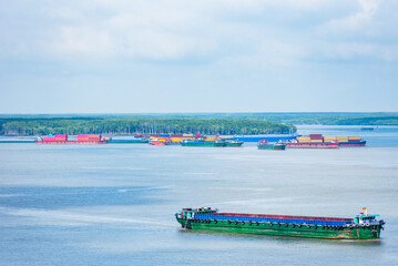 Obraz premium Barges and small boats navigate the flowing waters of the Mekong River in Vietnam, with a backdrop of lush tropical forest. 